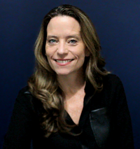 A woman with long wavy light brown hair, wearing a black blouse with a leather pocket detail, smiling against a solid dark blue background